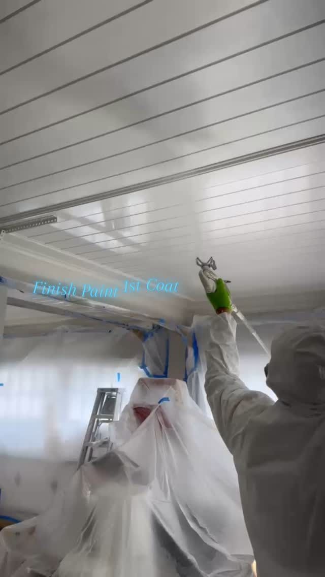 People in protective suits spray-clean a white ceiling in a bright room.