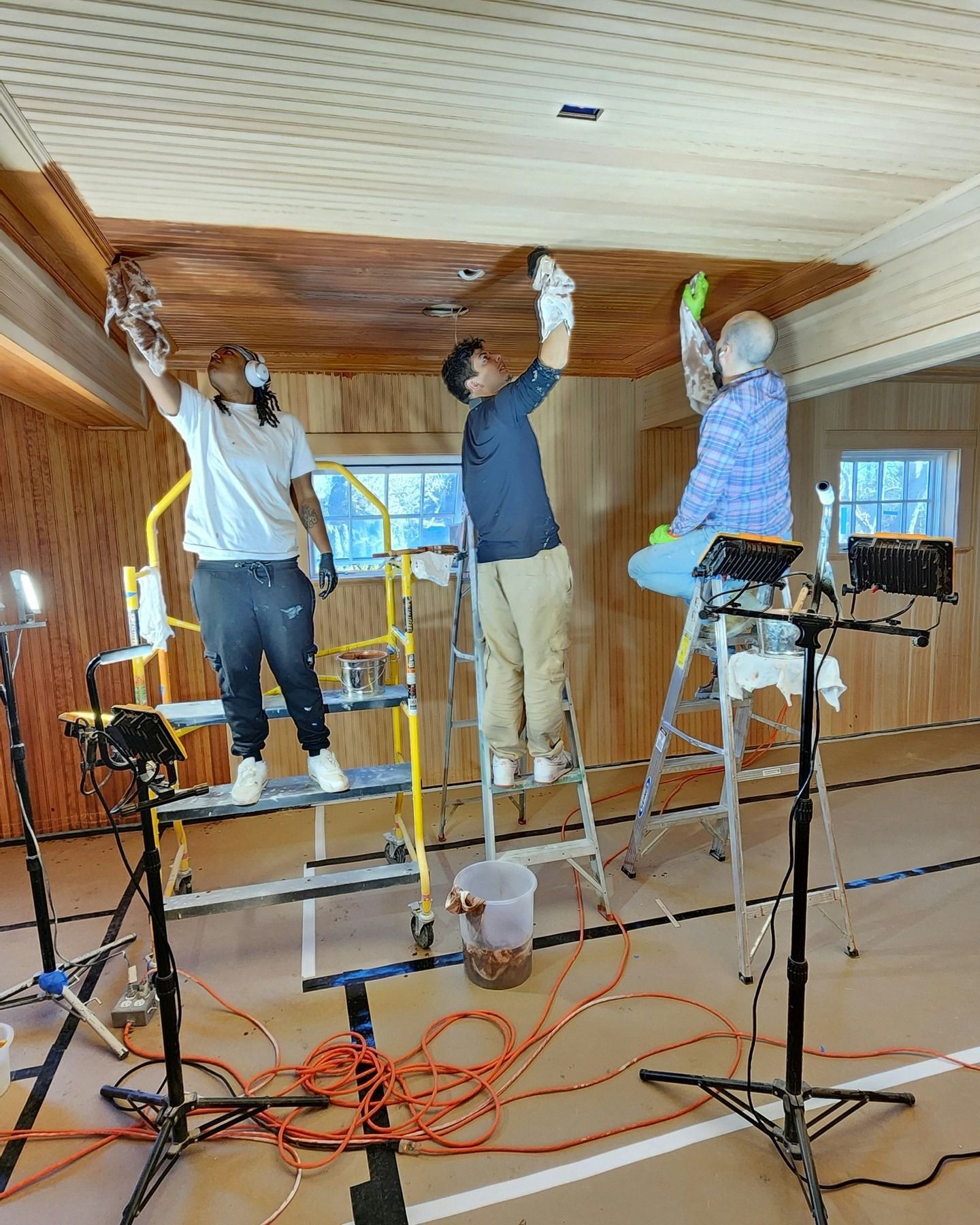 Three workers on ladders painting a wood-paneled ceiling in a room with equipment and extension cords.