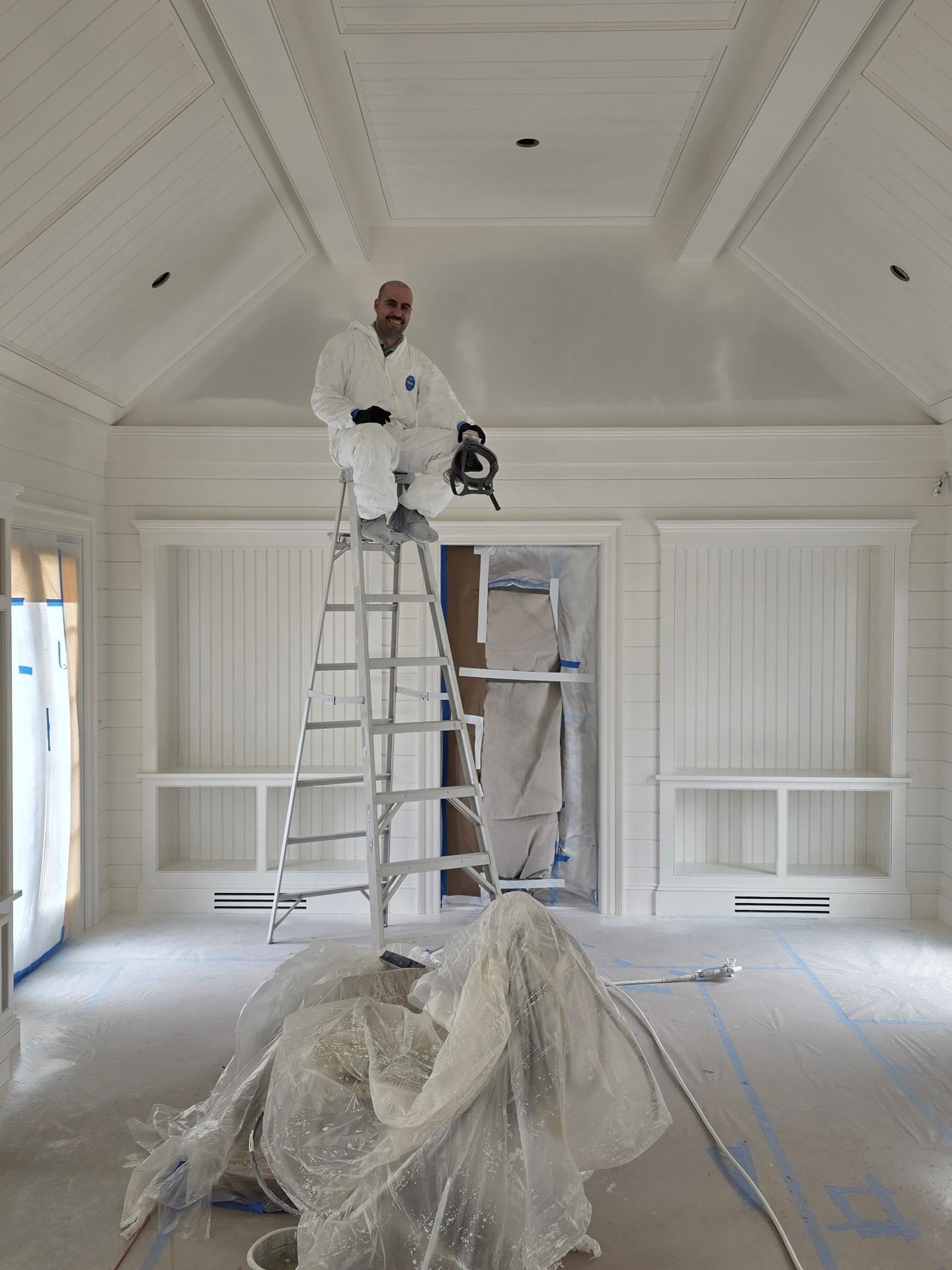 Painter on a ladder painting a white room with vaulted ceiling, plastic sheeting protecting the floor.