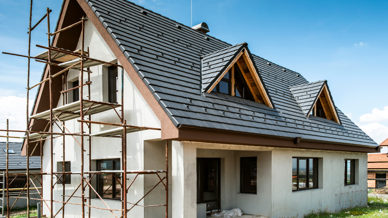 House under construction with scaffolding, gray roof, and triangular dormers against a blue sky.