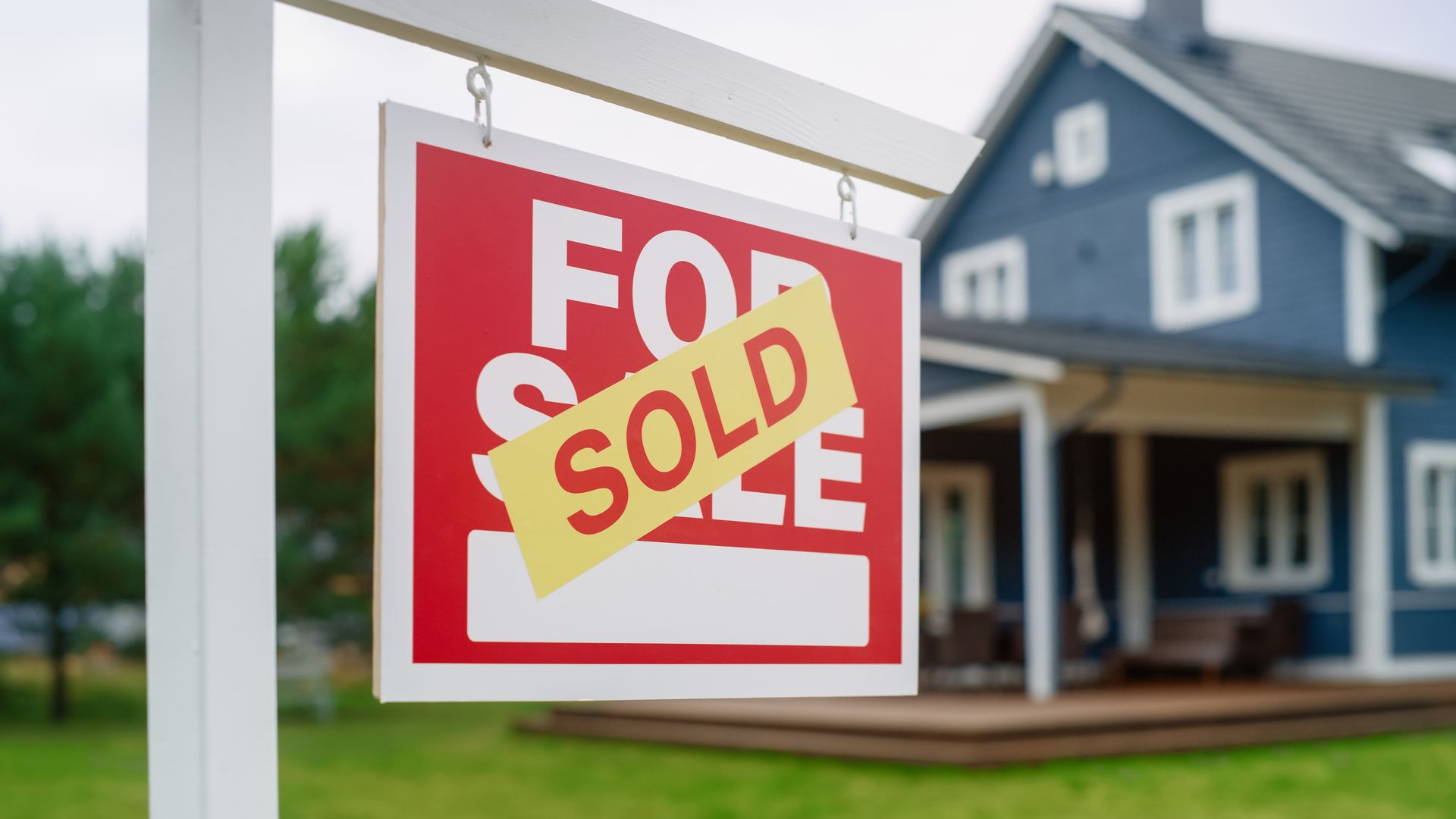 A for sale sign is hanging in front of a blue house.