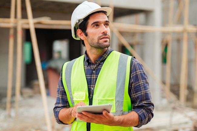 A construction worker wearing a hard hat and safety vest is holding a tablet.