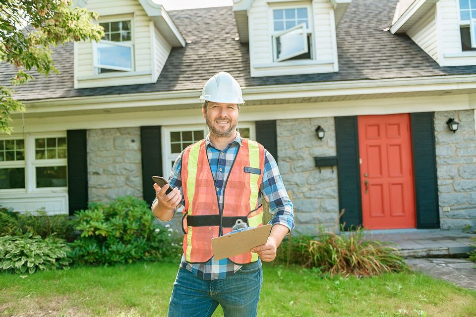 A construction worker is standing in front of a house holding a clipboard and a cell phone.