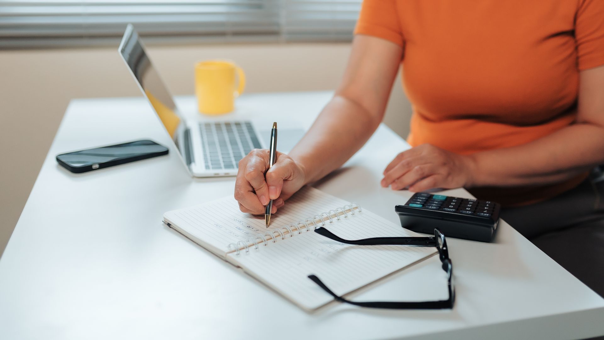 Person in orange top writing in a spiral notebook at a desk with a laptop, phone, and calculator.