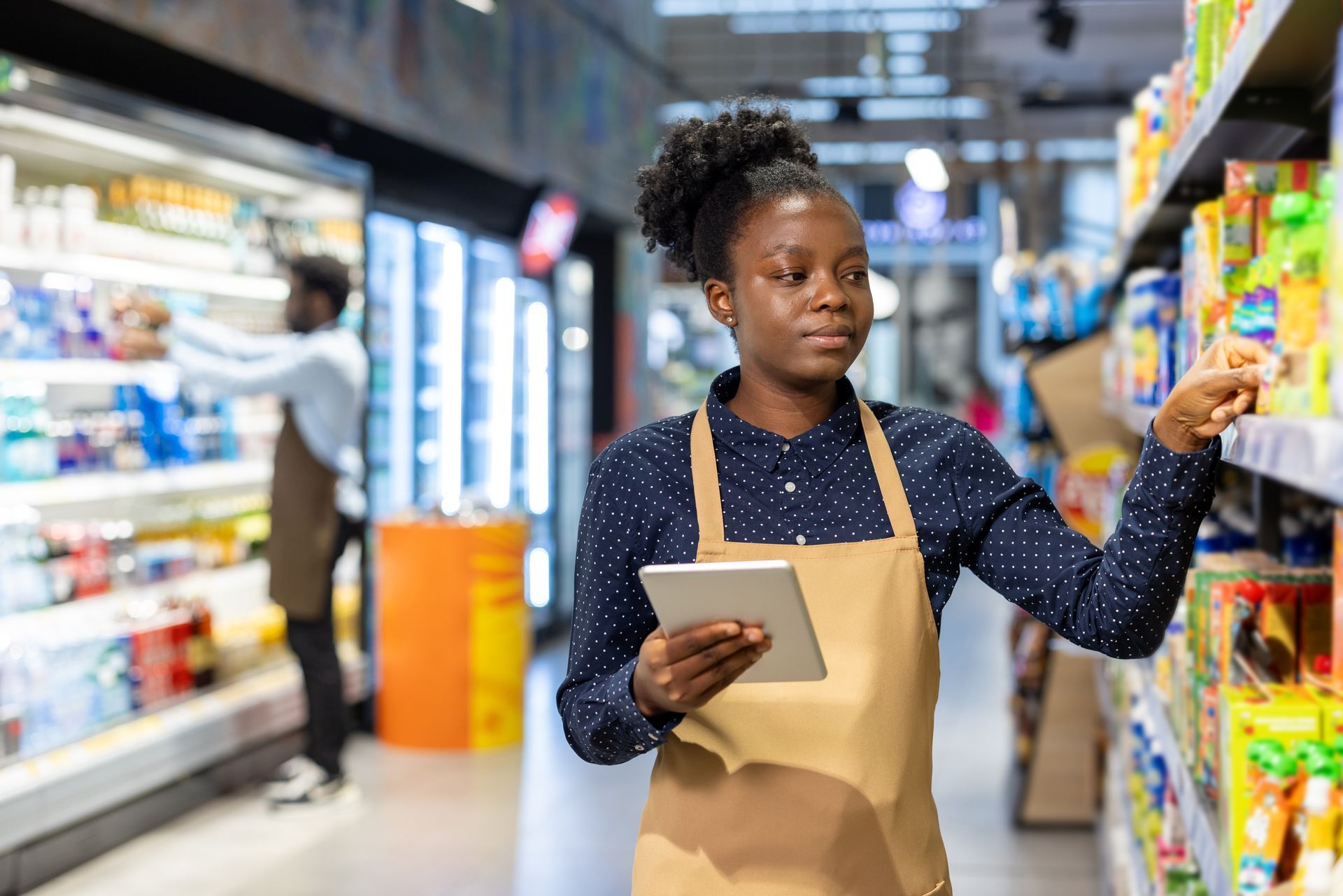 Woman in apron using a tablet to stock shelves in a grocery store. Another worker in the background.