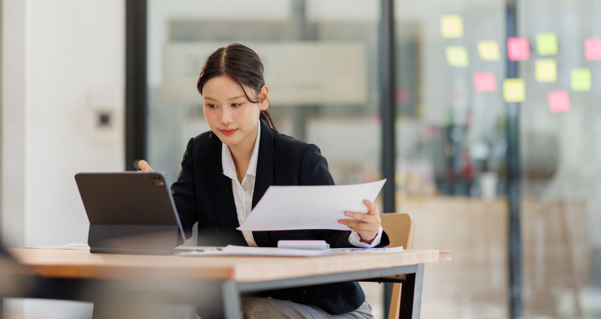 Woman in a blazer working at a desk with a tablet and papers in a modern office.