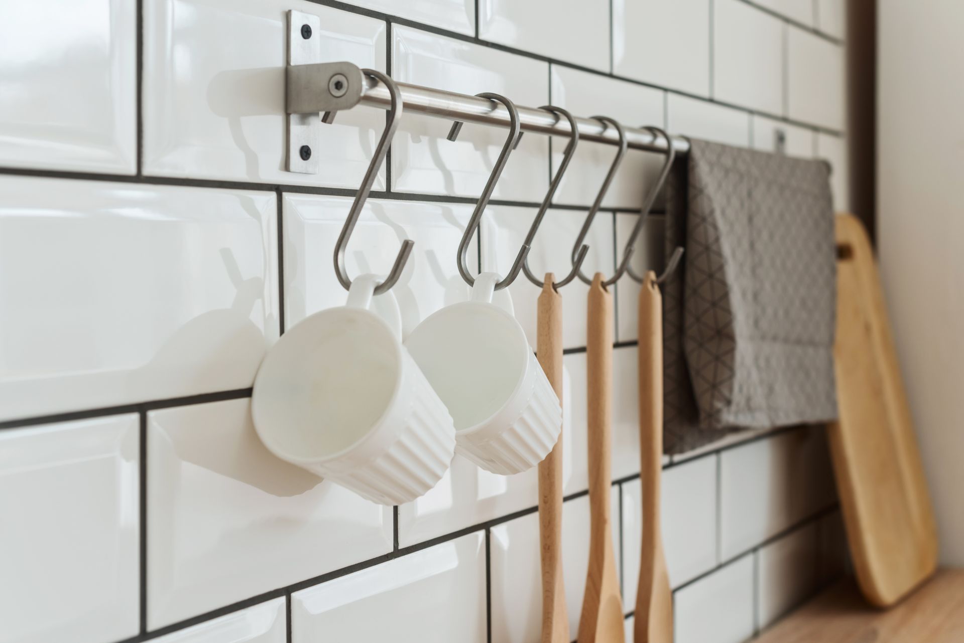 White ceramic mugs and wooden utensils hanging from an S-hook rack on a tiled kitchen wall.