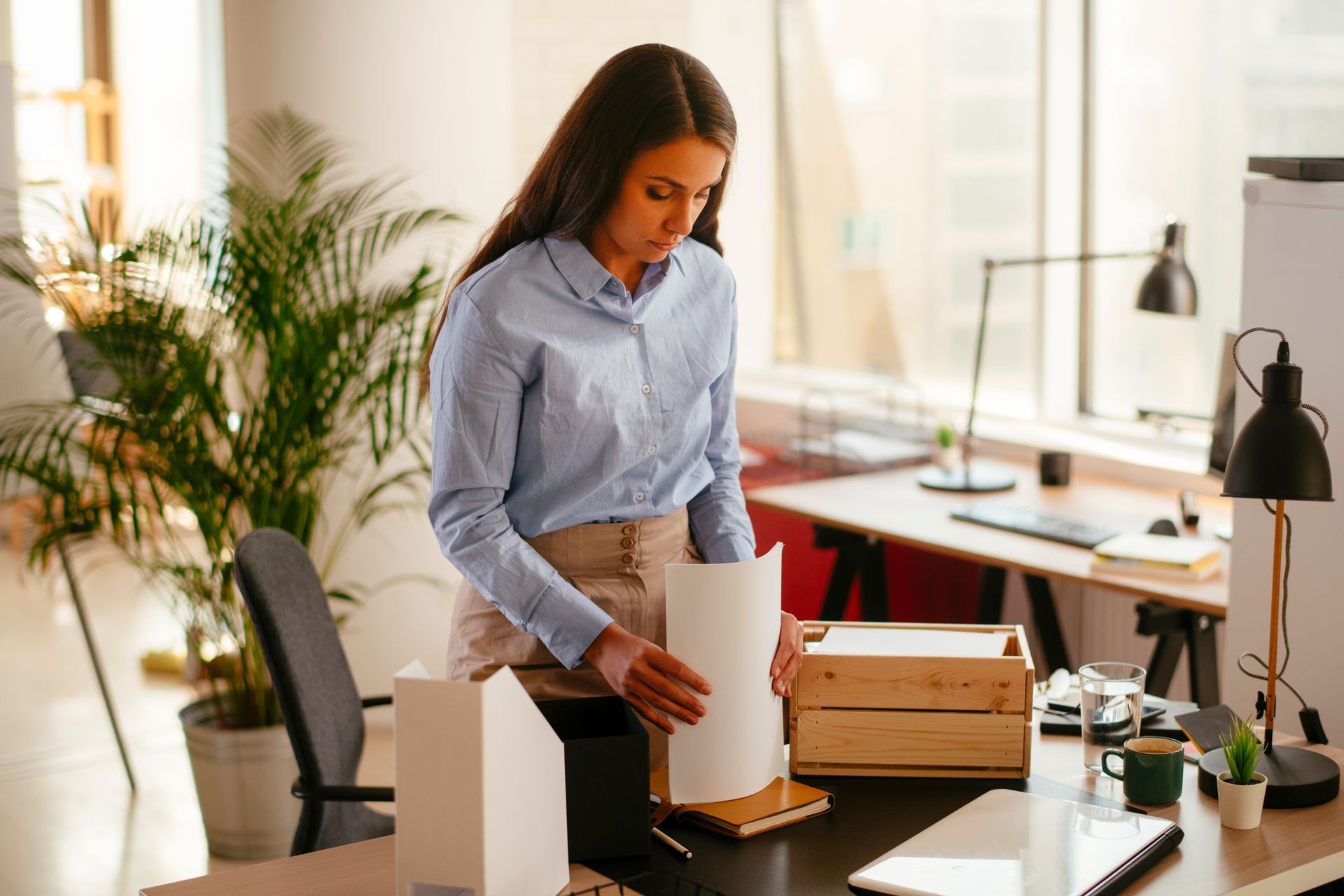 Woman in blue shirt arranging papers on a desk in an office setting.