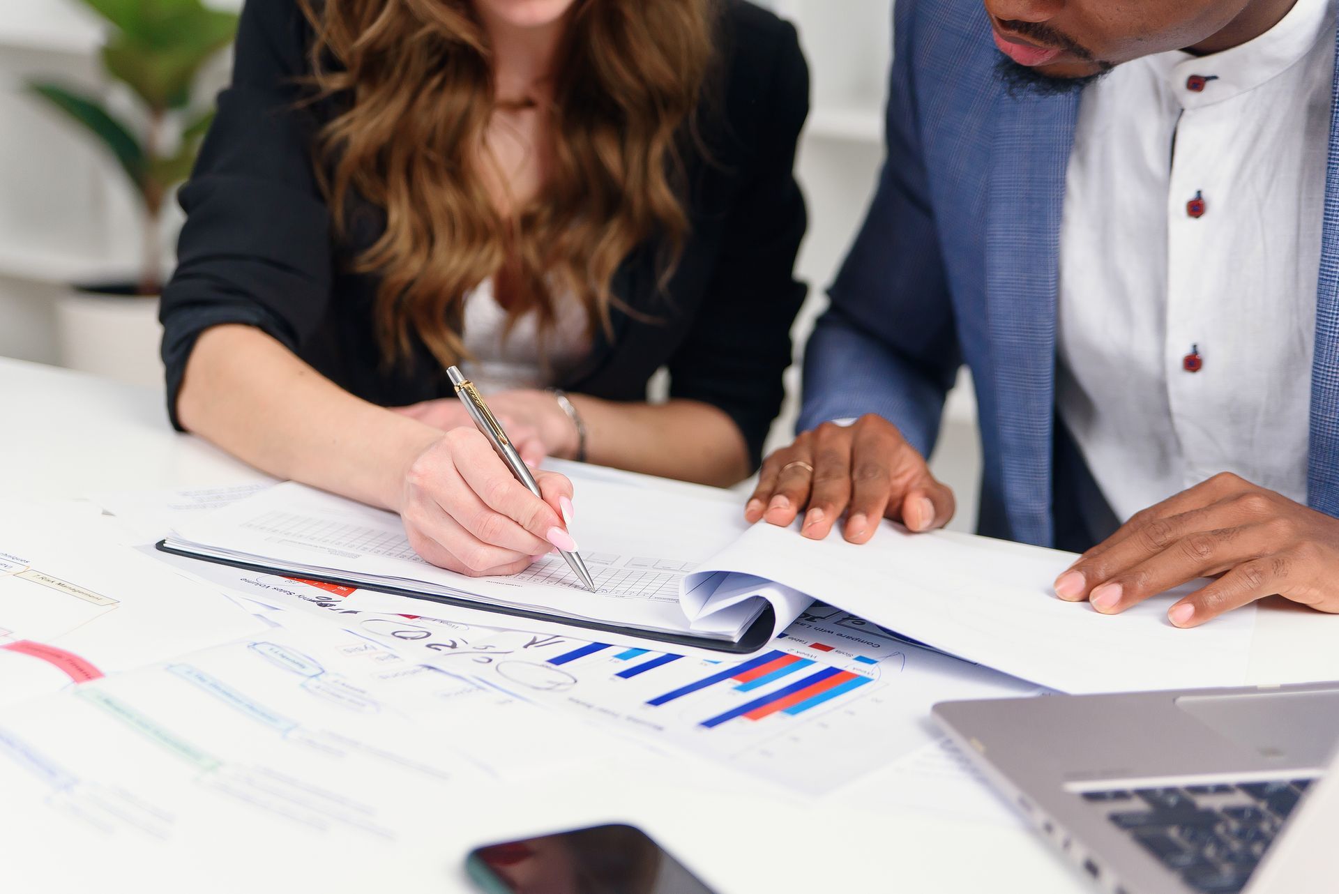 Two people reviewing documents at a white table, one writing with a pen, graphs, and laptop. Two people reviewing documents at a white table, one writing with a pen, graphs, and laptop.