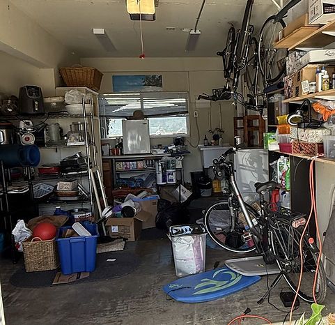 Cluttered garage interior: bikes, shelving with items, boxes, a surfboard, and a ladder.