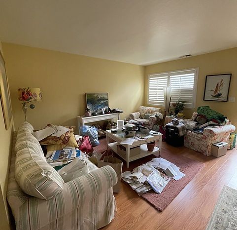 Cluttered living room: couch, chairs, coffee table, and various items strewn about; yellow walls and window shutters.