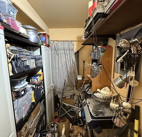 Cluttered room: shelves and surfaces overflow with items, narrow passageway, neutral walls, curtained window.