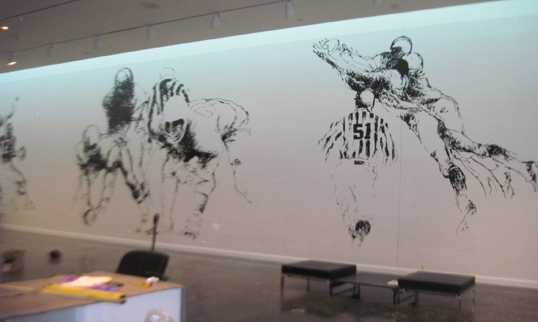 Black and white mural of stylized figures, possibly athletes, on a white wall in a modern room; includes a desk and benches.