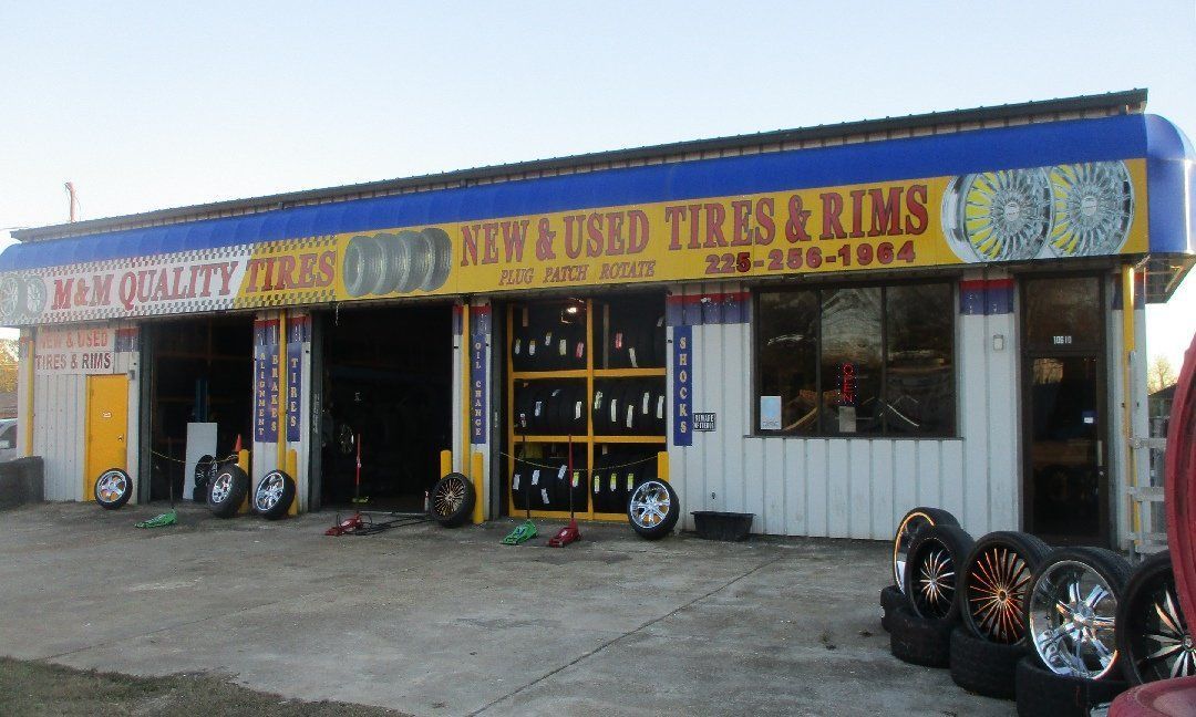 Exterior of M&M Quality Tires shop displaying tires and rims for sale. Yellow and blue awning, open garage bays, and tires stacked outside.
