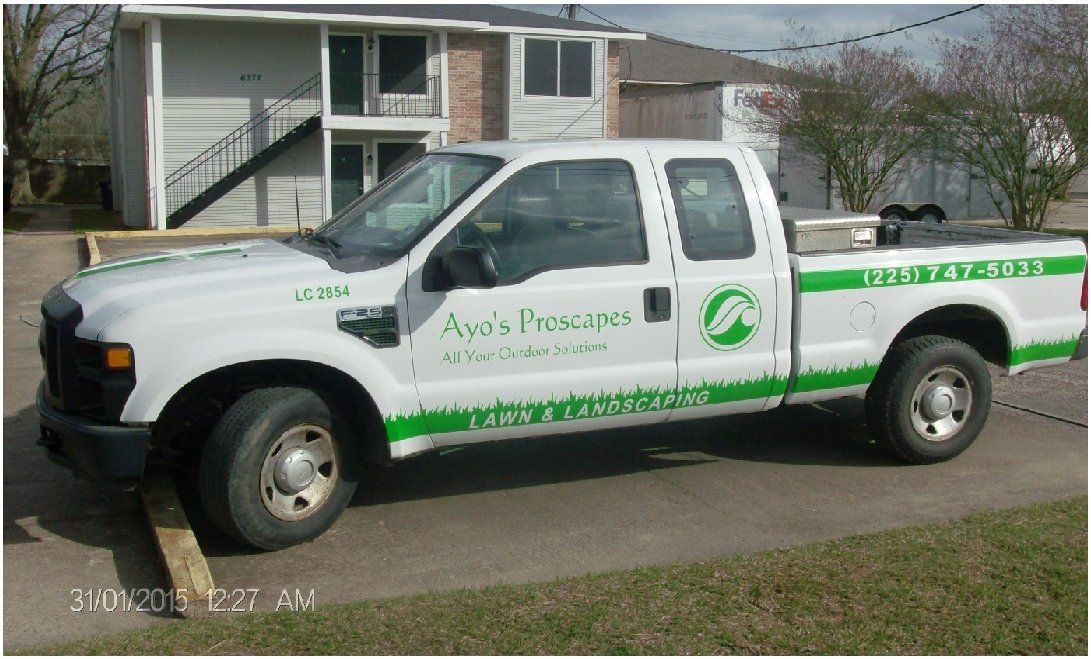 White pickup truck with green landscaping company logo parked in front of a multi-story building.