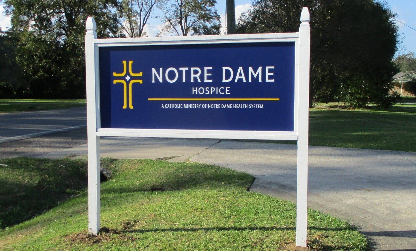 Sign for Notre Dame Hospice, featuring a gold cross on a navy blue background, set in front of green grass.