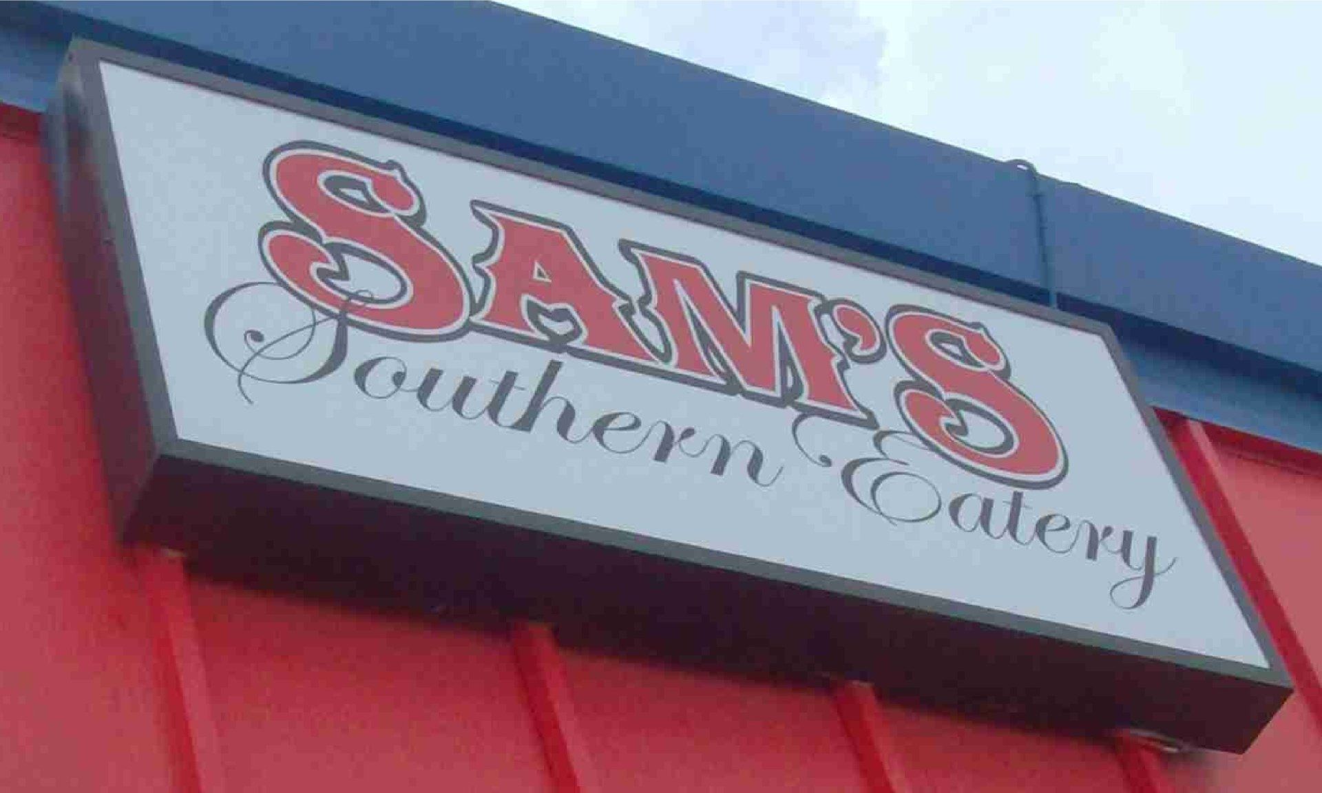 Sam's Southern Eatery sign with red lettering and a white background. The sign hangs against a red and blue building.