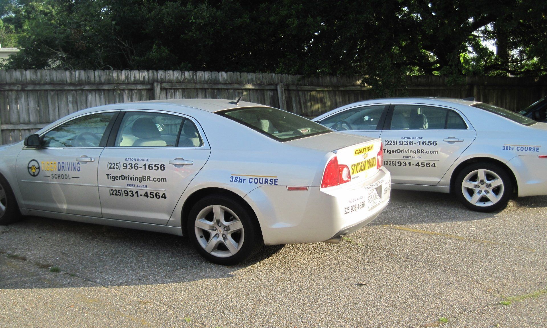 Two silver cars parked side-by-side, likely taxis, with business logos on the doors, in front of a wooden fence.