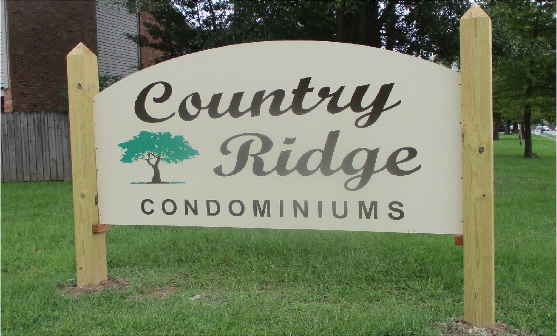 Country Ridge Condominiums sign, beige with brown text, green tree graphic. Mounted on wooden posts in a grassy area.