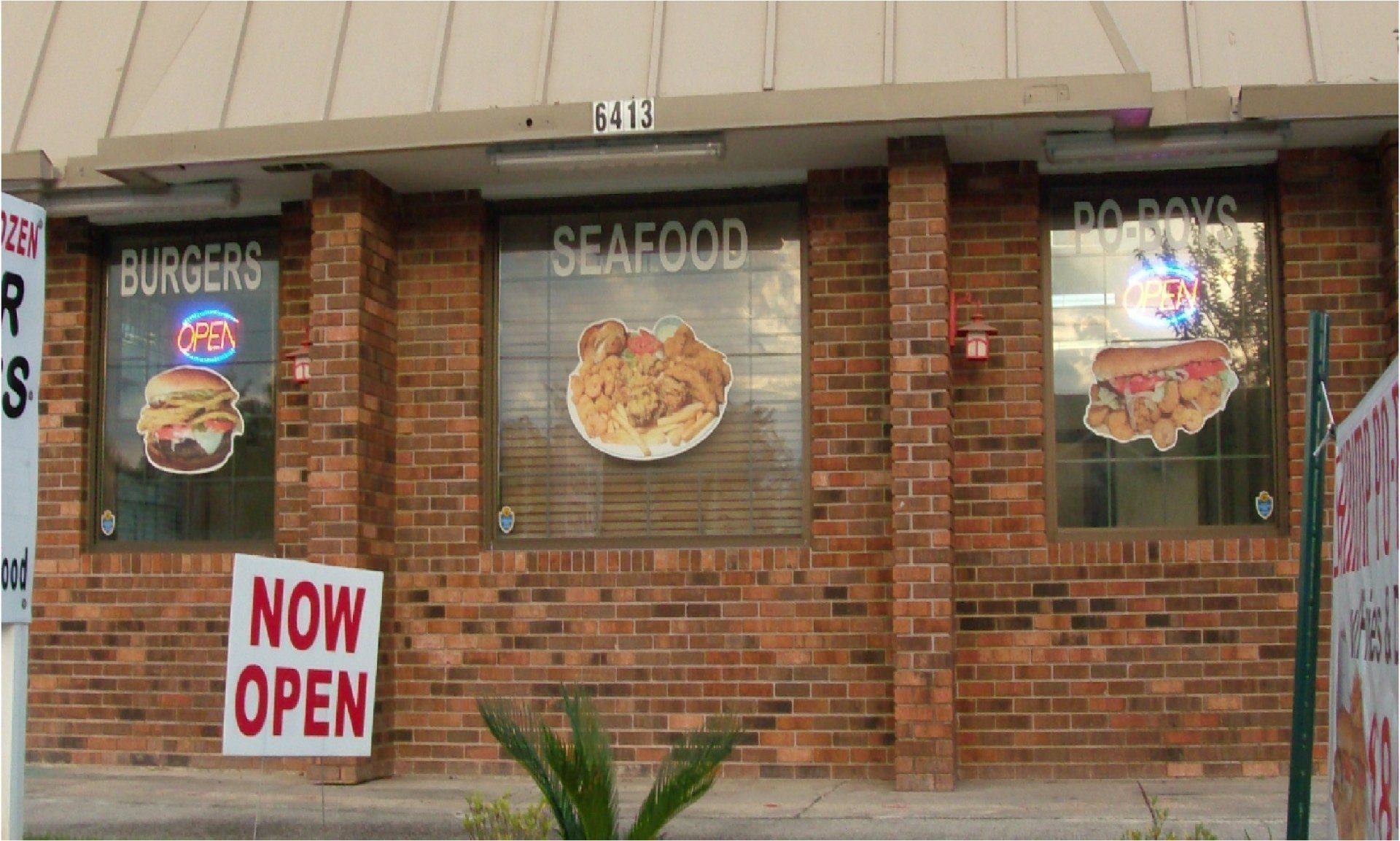 A brick building with windows displaying food options: burgers, seafood, and po' boys. 