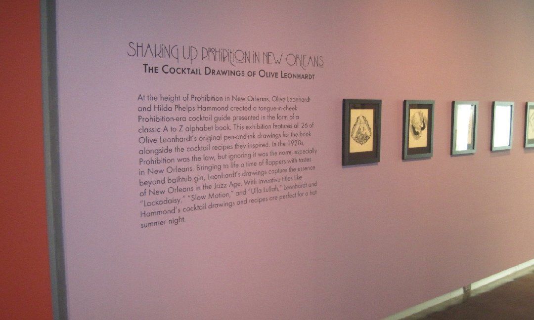 A museum wall with text and framed art. The wall is a muted purple with text on the left. Several framed pieces of art hang on the right.