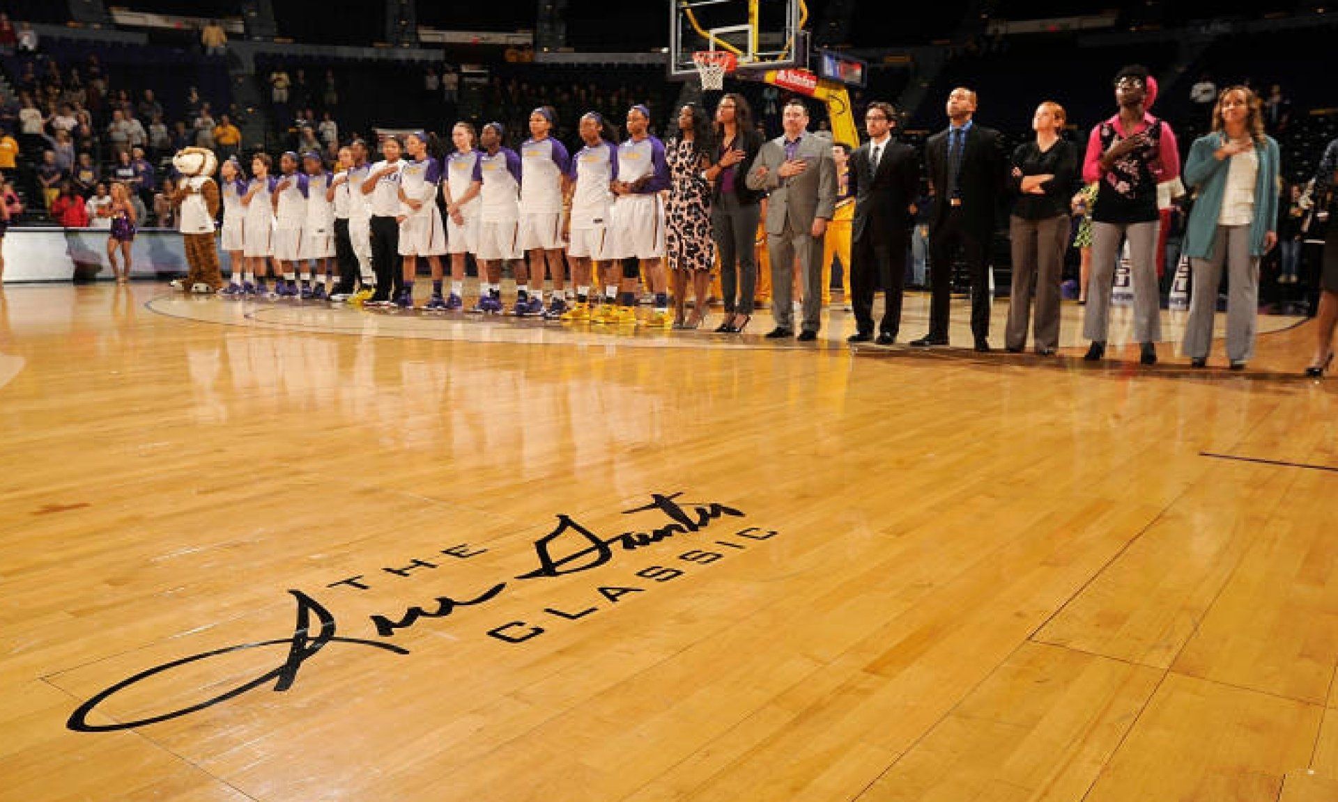 A basketball court with players, officials, and others standing for the national anthem. A large signature on the floor reads 