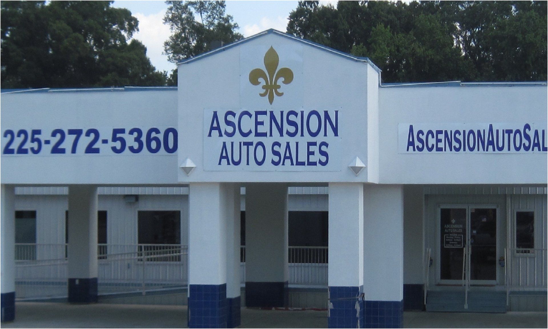 Ascension Auto Sales storefront with blue accents and a fleur-de-lis logo, in front of a cloudy sky.