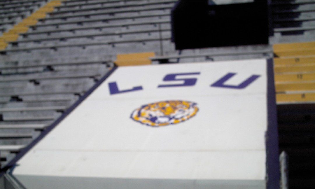 LSU logo and lettering in purple and gold, painted on a white wall inside a stadium. The background shows rows of empty seats.