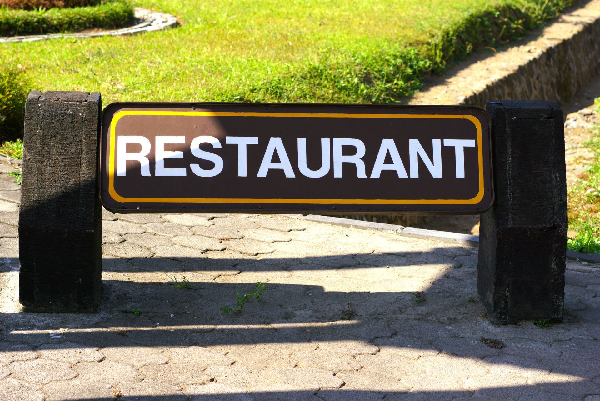 Restaurant sign with brown background and gold border, white text, supported by dark grey pillars, set in a paved area with grass.