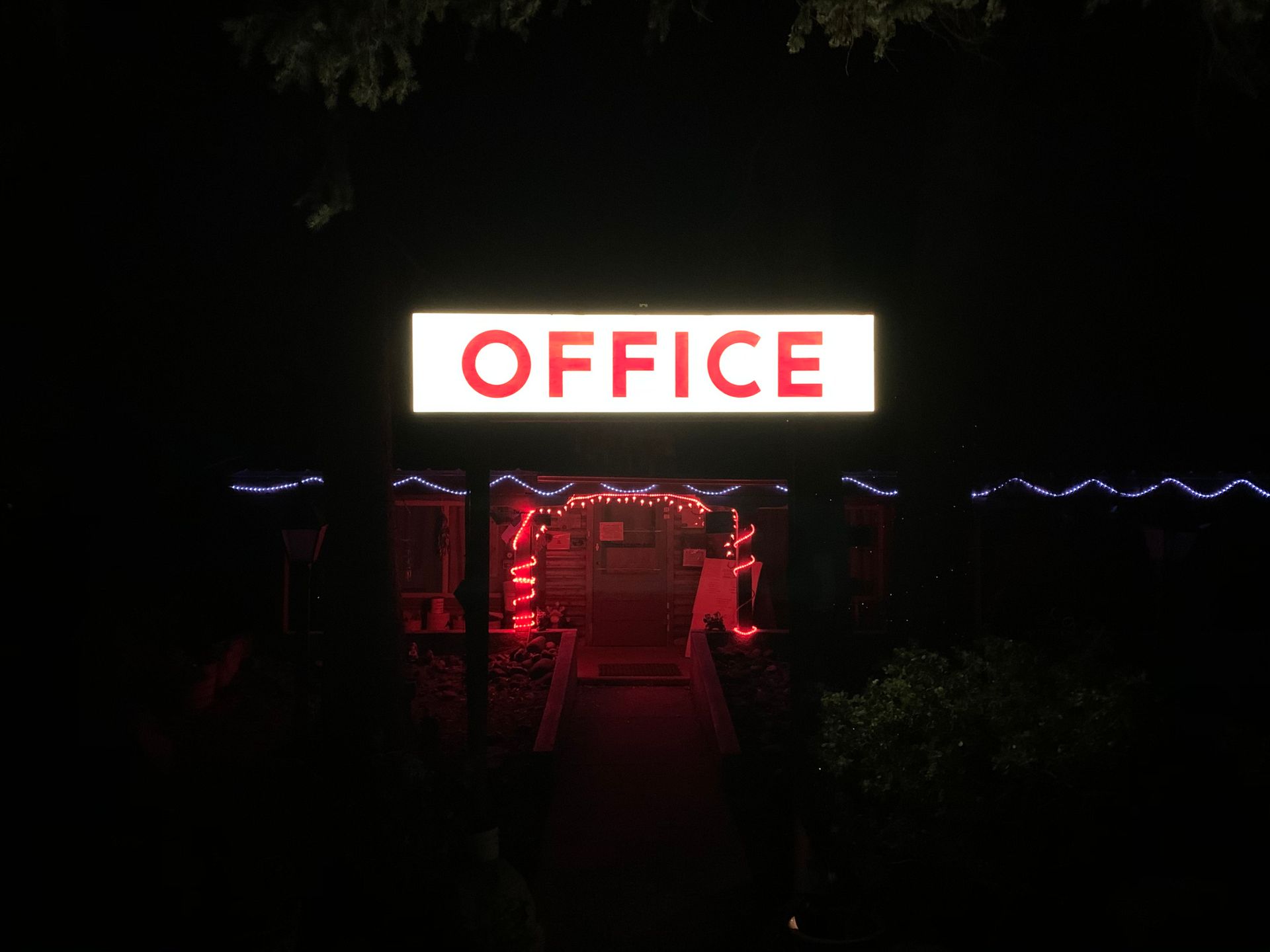 Office sign lit up in red, above a doorway at night. Decorated with red and blue lights.