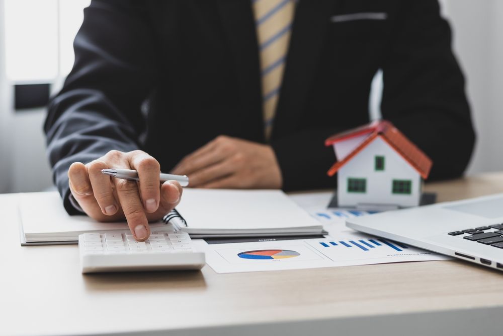 Businessman calculating his house value using a calculator and pen on a desk with papers and a laptop nearby.