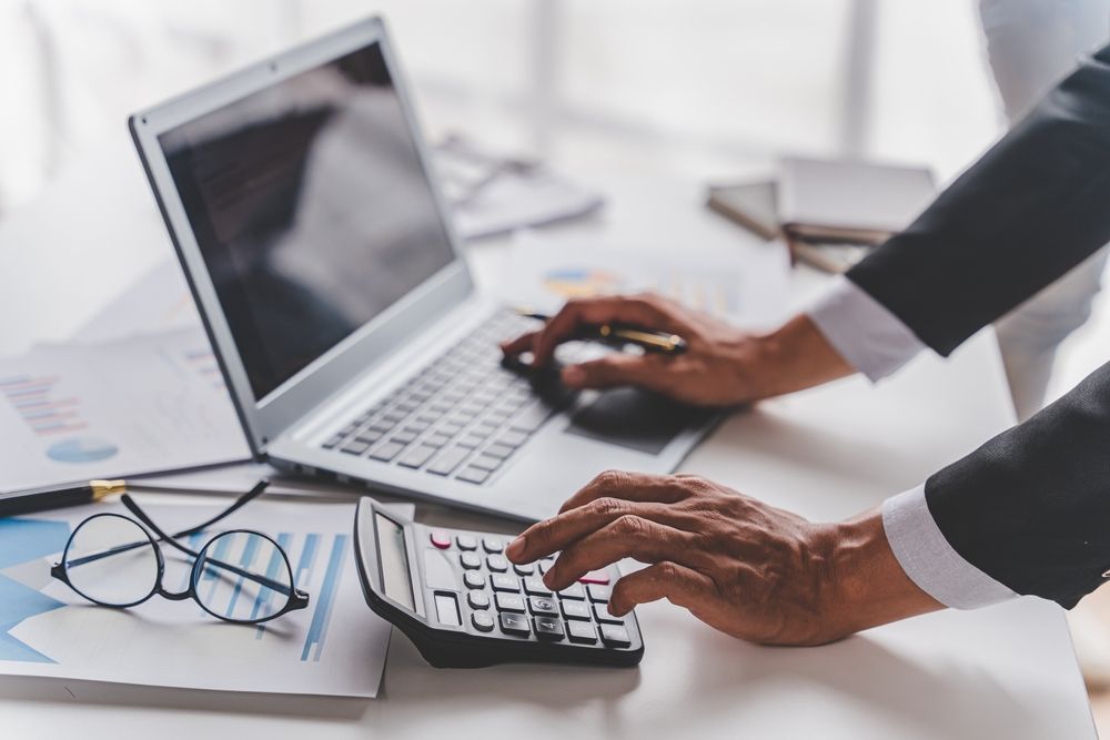 A person in a suit calculates using a calculator while working on a laptop at a desk.