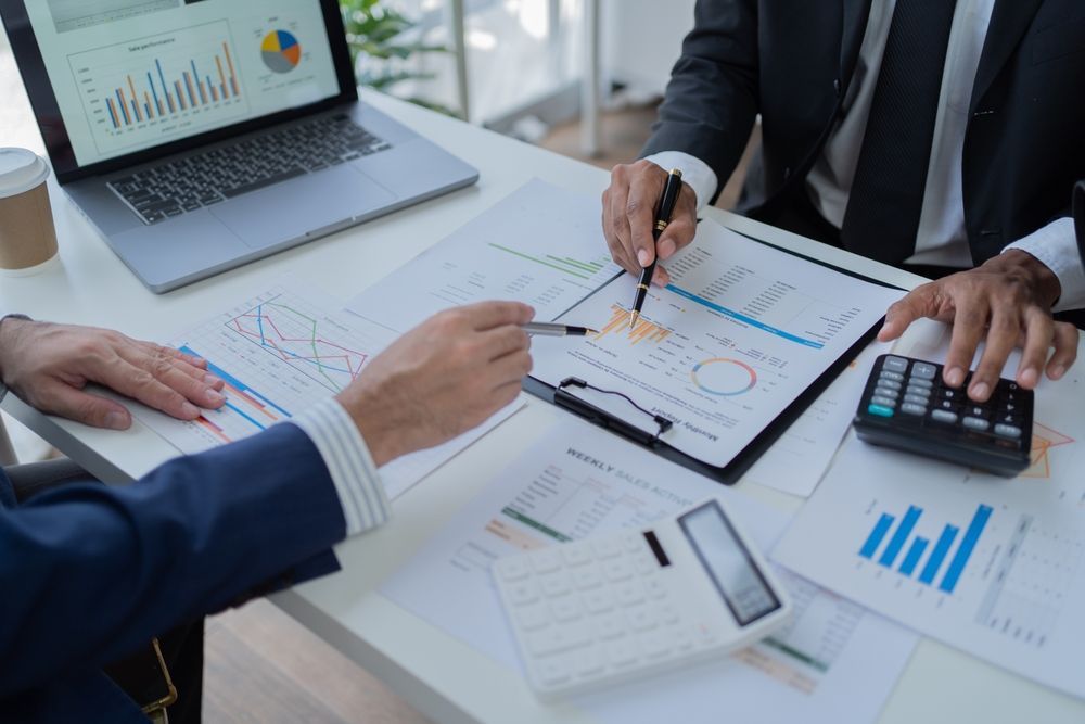 Business professionals collaborating on a laptop, surrounded by papers and documents in a modern office setting.