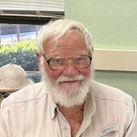 Smiling man with white hair and beard wearing glasses in a light-colored shirt, seated indoors near a window