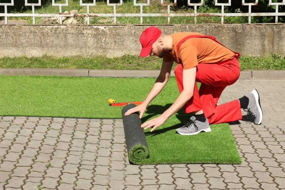 Worker in red kneels on paving stones, rolling out green artificial turf on a lawn edge.