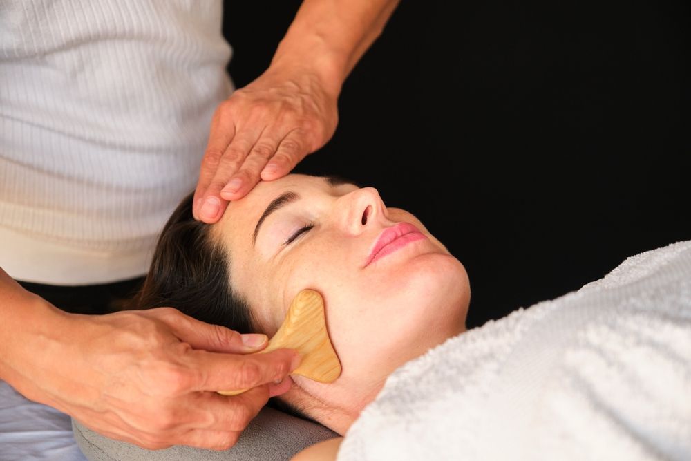 Woman receiving facial Maderotherapy treatment; hands on forehead and cheek, jade tool visible.