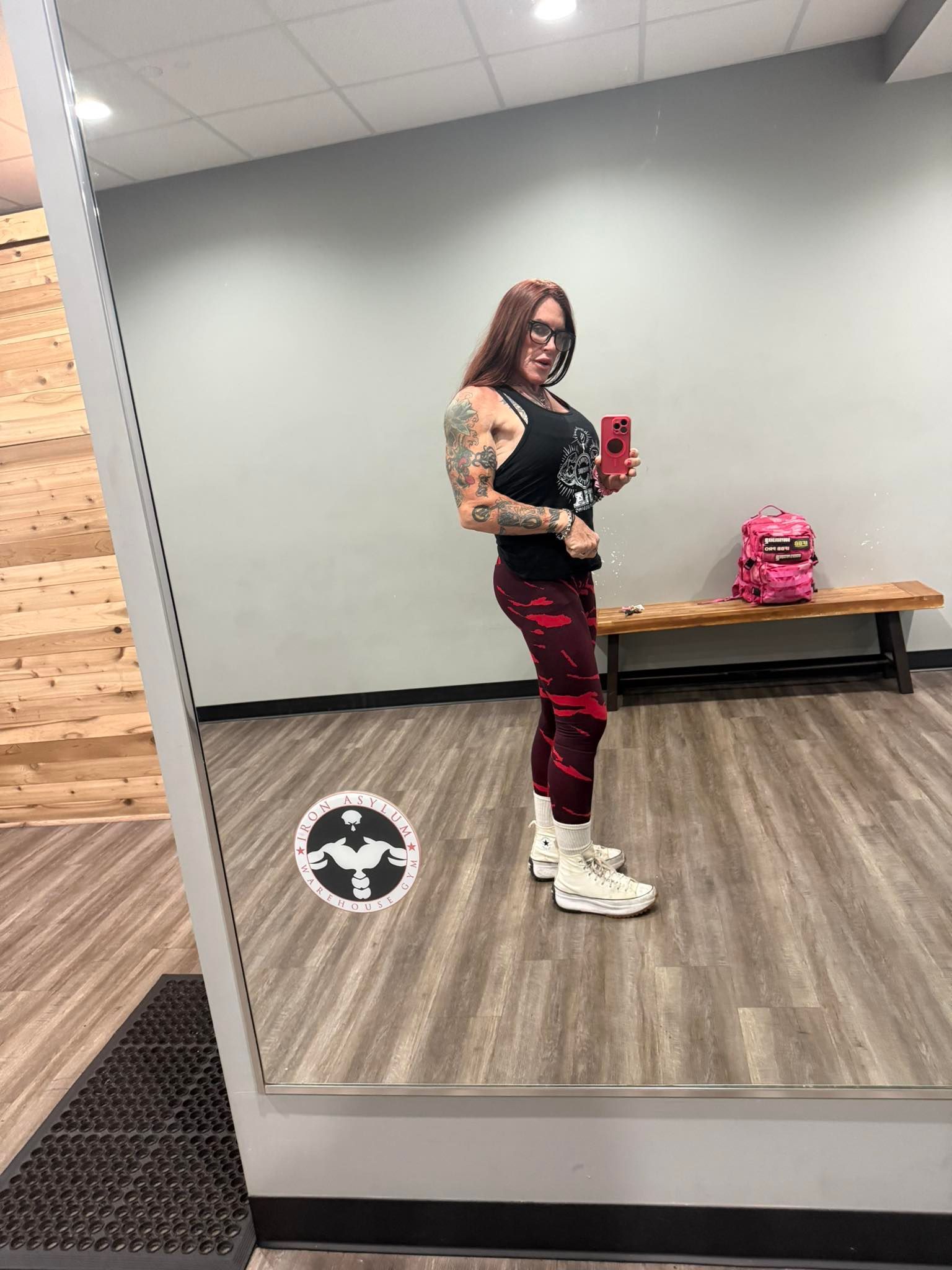 Woman in gym clothes poses in front of a mirror, holding a phone. Workout setting.