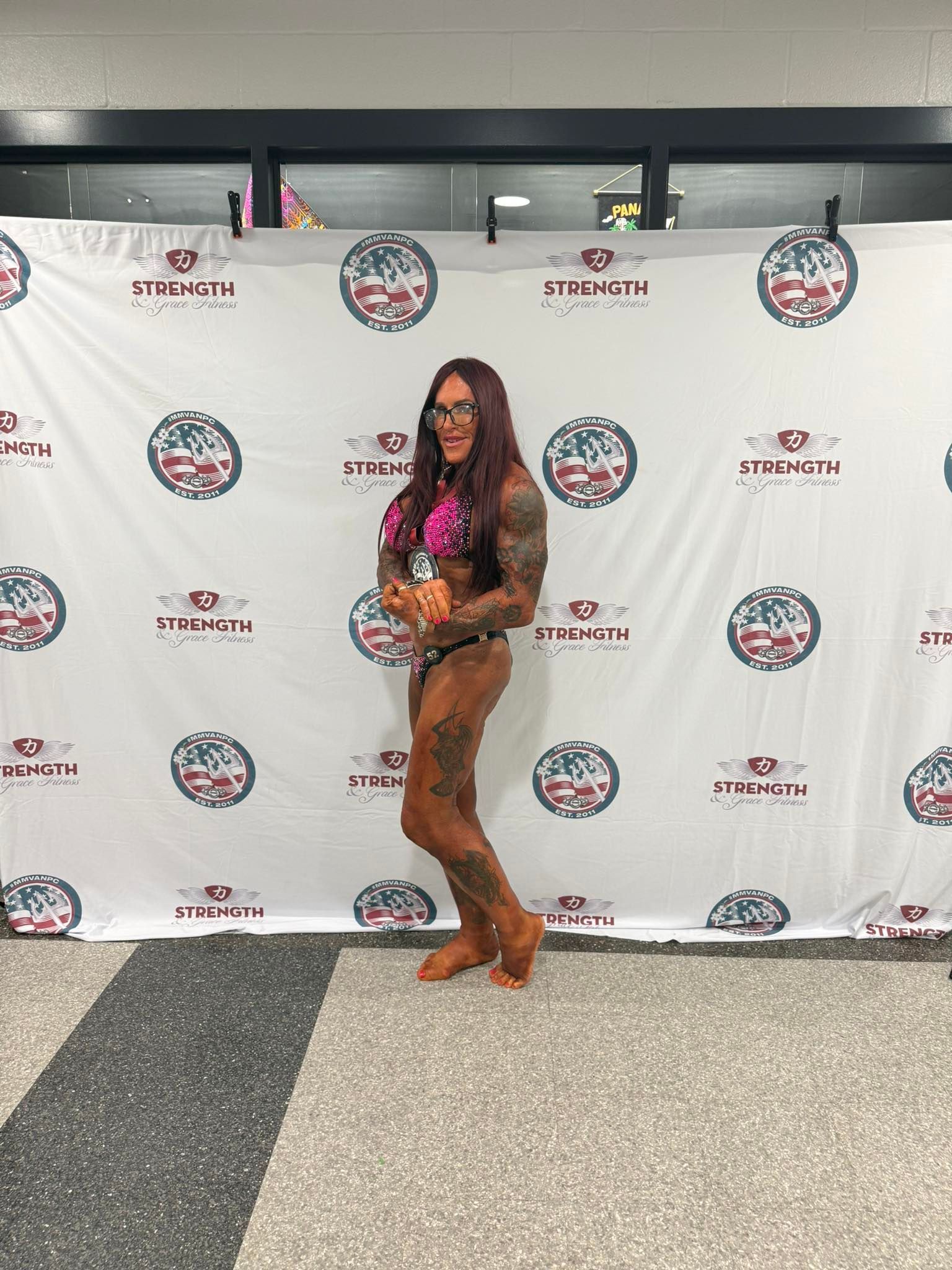 Woman in bodybuilding pose with tattoos, wearing a bikini, in front of a banner.