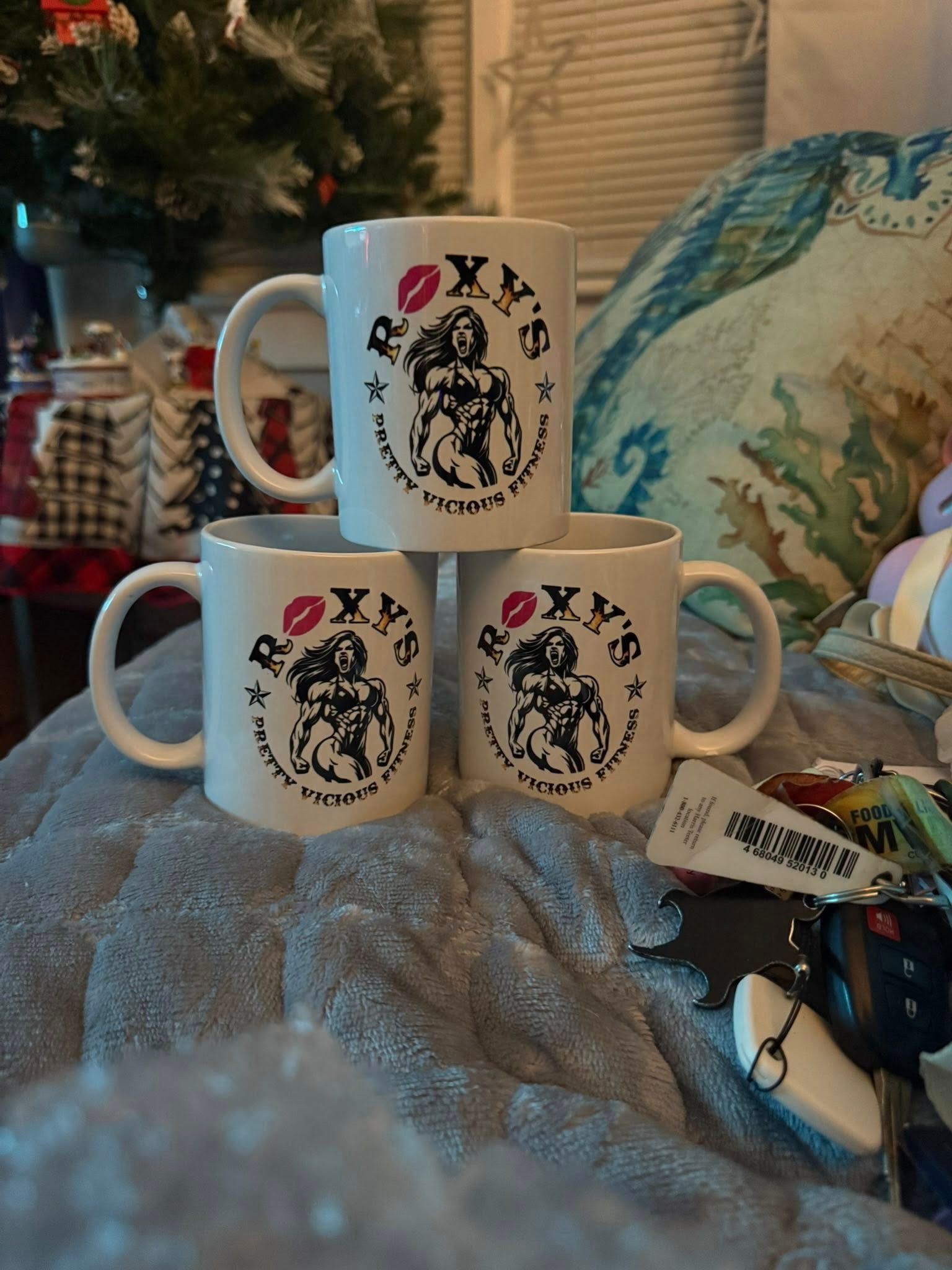 Three white mugs stacked, each with a centered design and red lip print. The mugs sit on a gray fuzzy surface.