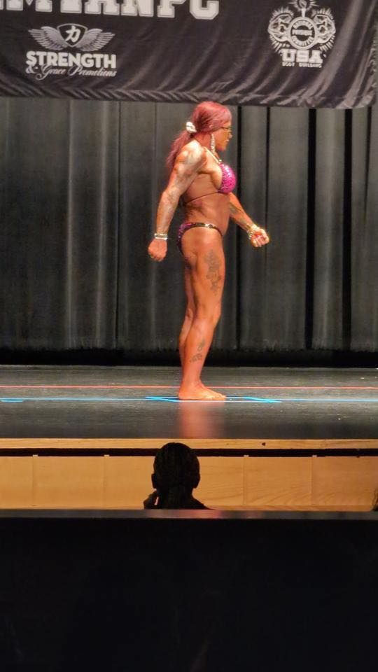 Bodybuilder in a pink bikini poses onstage, tanned skin, arms flexed, black backdrop, contest logo visible.