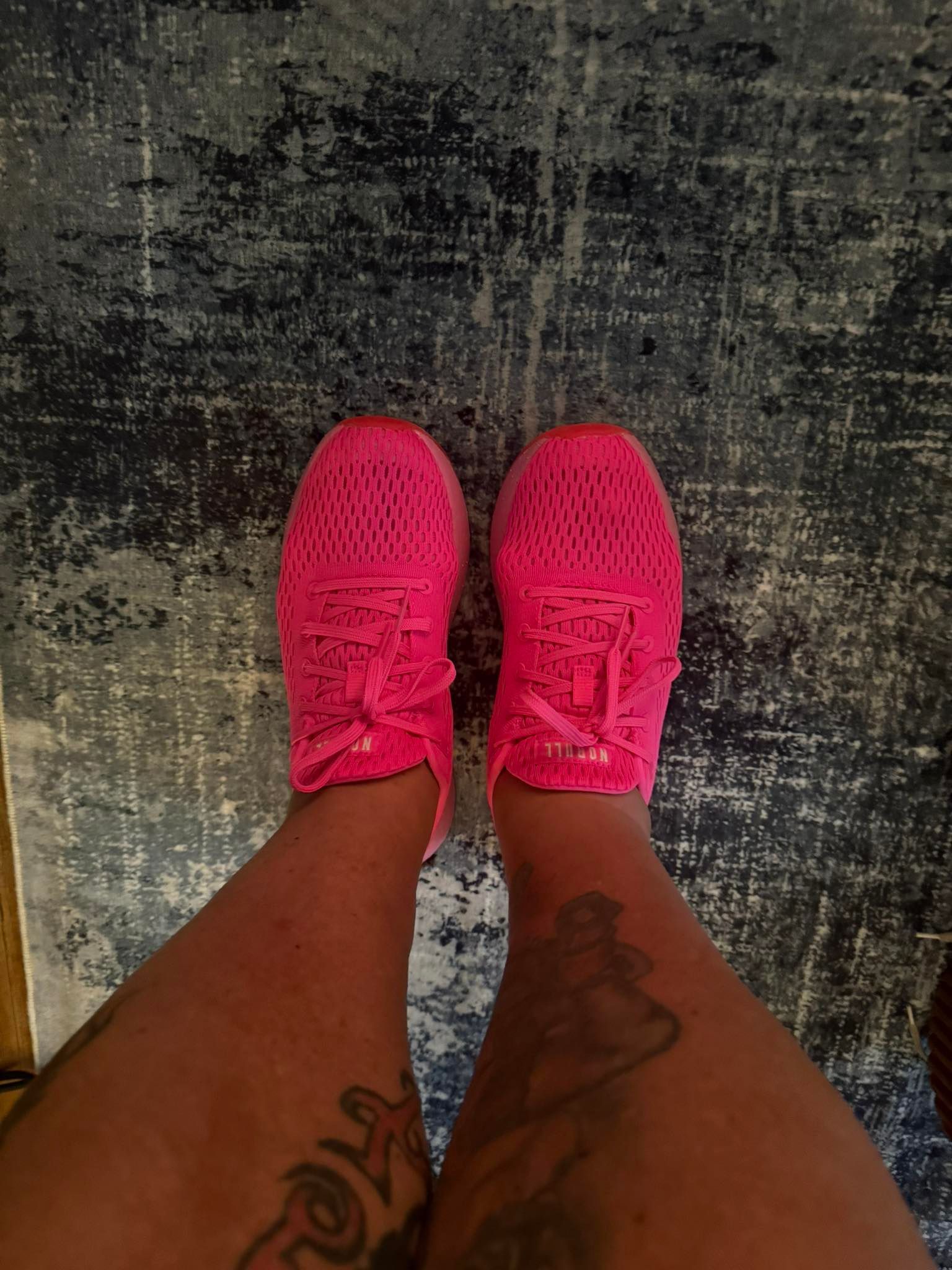 Person's legs and bright pink sneakers on a dark patterned rug; visible leg tattoos.