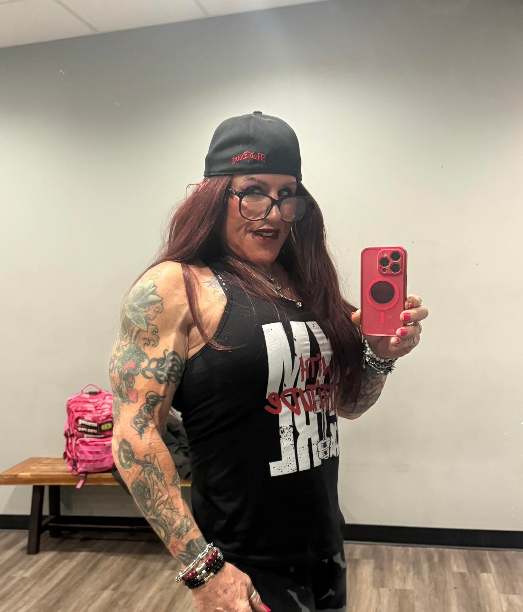 Woman in a gym, flexes arm, holding a phone, wearing a hat, glasses, and a tank top.