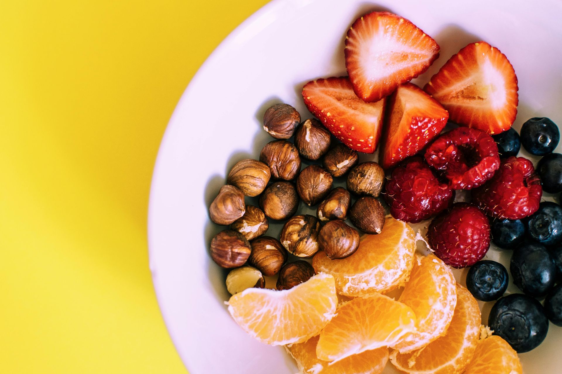 Close-up of a white bowl with colorful food: strawberries, blueberries, hazelnuts, raspberries, and orange slices, on a yellow surface.
