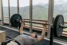 Man bench presses barbell in gym with mountain view.