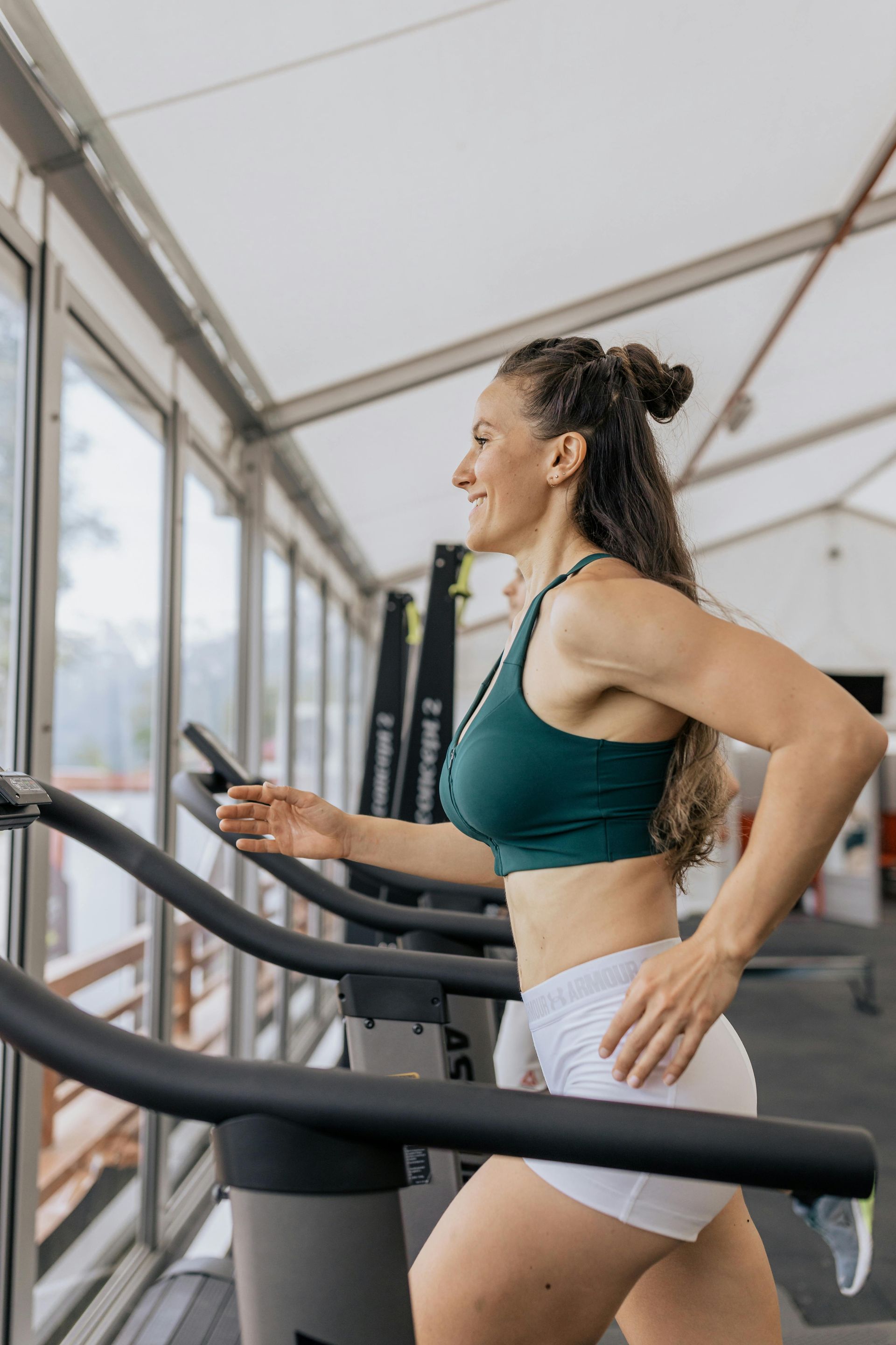 Woman in a green sports bra and white shorts running on a treadmill at a gym, smiling.
