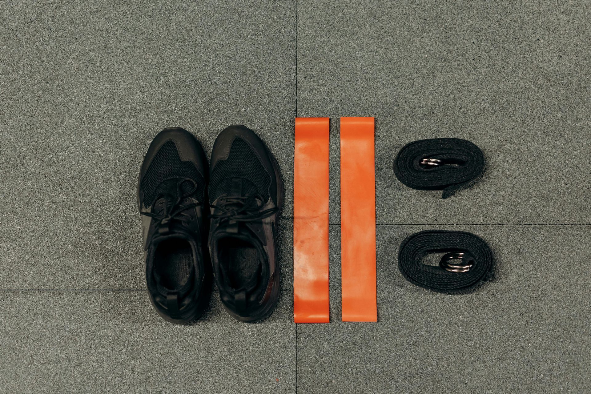Black sneakers, orange resistance bands, and black ankle straps on a gray floor.