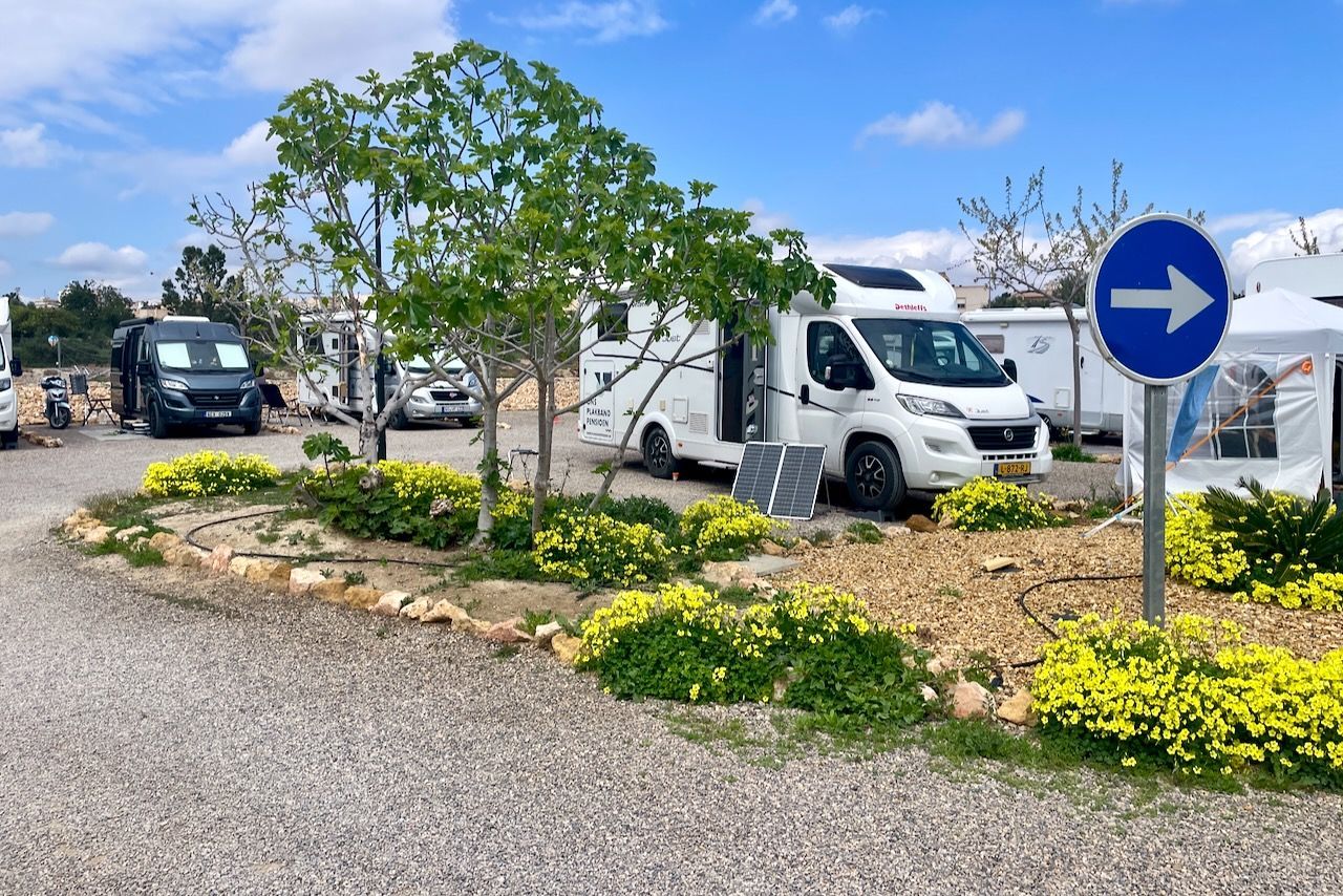 Recreatieve voertuigen geparkeerd op een grindterrein met een blauw richtingaanwijzerbord en bloeiende planten op de voorgrond.