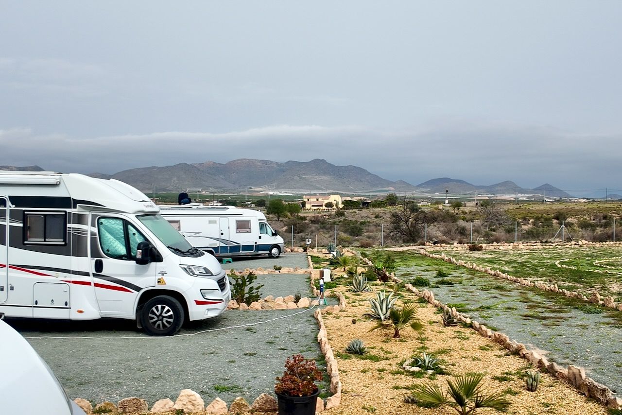 Twee campers staan ​​geparkeerd op een camping, met uitzicht op een landschap van bergen en dorre vegetatie onder een bewolkte hemel.