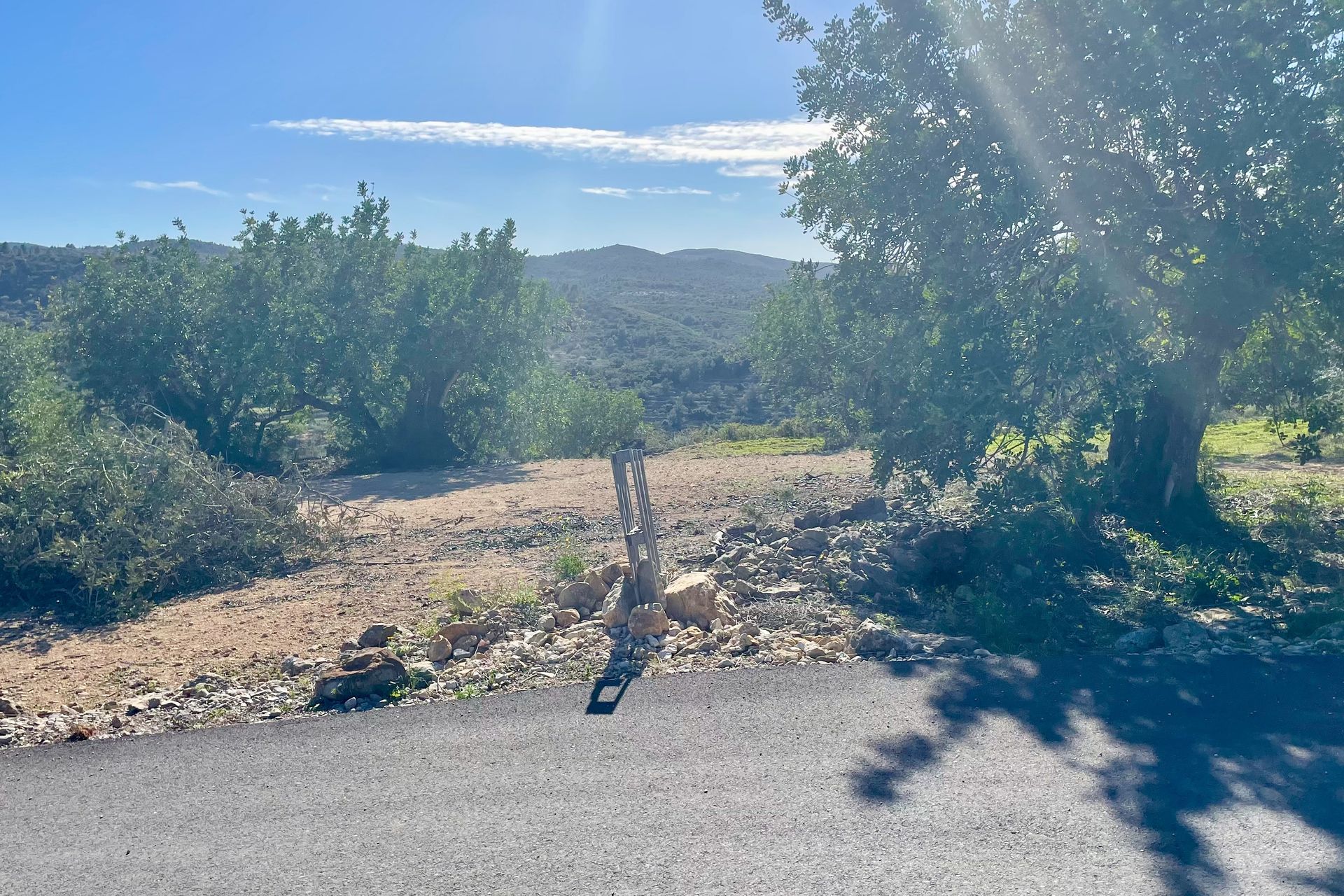 Een weg die overgaat in een onverhard pad, met bomen aan weerszijden en heuvels op de achtergrond op een zonnige dag.