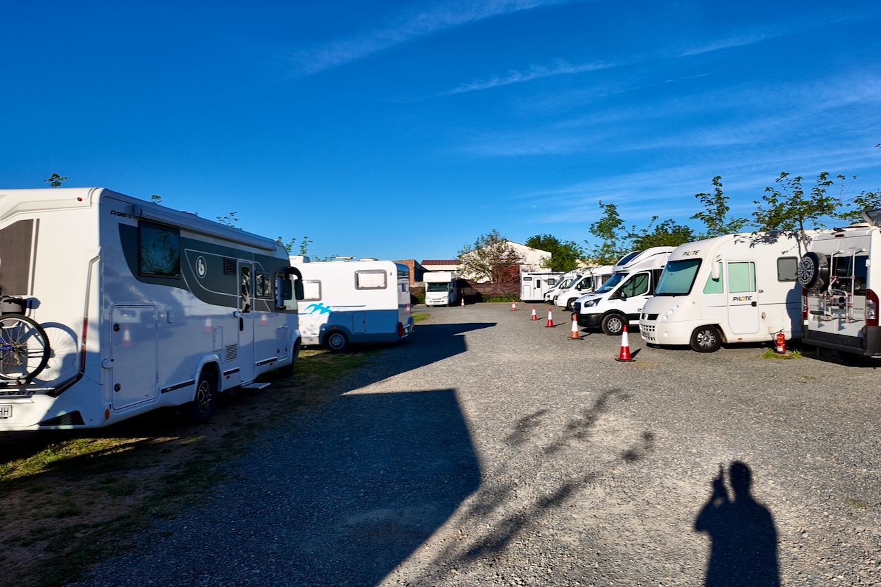 Een grindparkeerplaats vol met diverse geparkeerde campers en caravans onder een helderblauwe hemel.